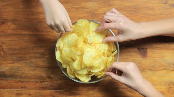 Drei Hände greifen in eine Schüssel Chips, die auf einem Tisch steht. | © AdobeStock-322465996