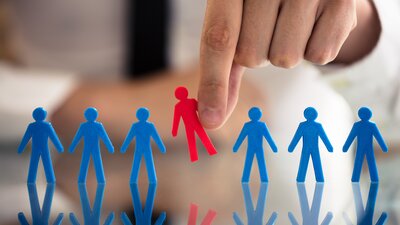 Eine rote Scherenschnittfigur wird aus einer Reihe von blauen herausgenommen (Symbolbild). | © AdobeStock_209445070