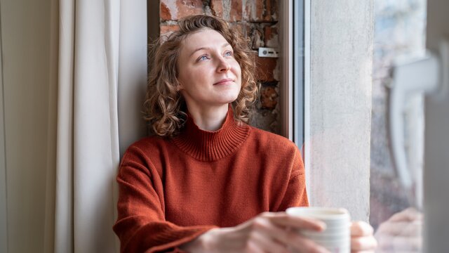 Eine Frau sitz am Fenster mit einer Tasse in der Hand und blickt in den Himmel. | © AdobeStock_1753411608