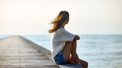 Eine junge Frau sitzt auf einem Steg und blickt aufs Meer. | © AdobeStock-117720676