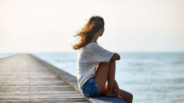 Eine junge Frau sitzt auf einem Steg und blickt aufs Meer. | © AdobeStock-117720676