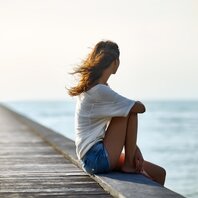 Eine junge Frau sitzt auf einem Steg und blickt aufs Meer. | © AdobeStock-117720676