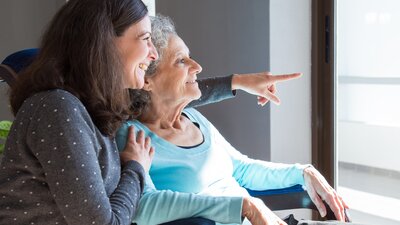 Eine jüngere Frau umarmt eine ältere Frau von der Seite - beide blicken aus einem Fenster.  | © AdobeStock-250496474