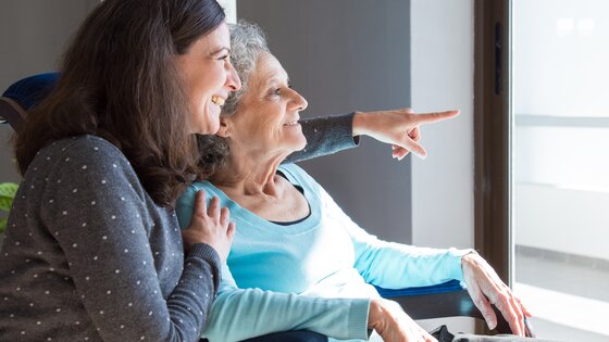 Eine jüngere Frau umarmt eine ältere Frau von der Seite - beide blicken aus einem Fenster.  | © AdobeStock-250496474