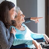 Eine jüngere Frau umarmt eine ältere Frau von der Seite - beide blicken aus einem Fenster.  | © AdobeStock-250496474