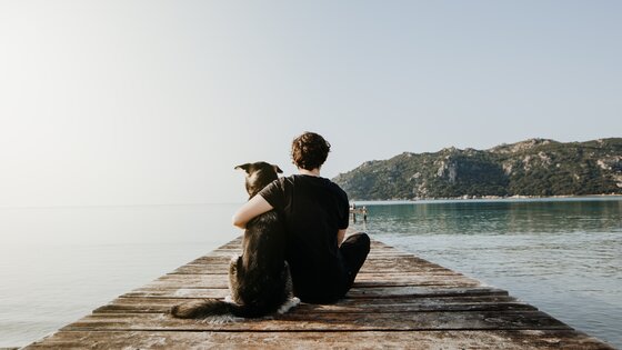 Ein Mann sitzt mit einem Hund auf einem Steg und schaut aufs Wasser. | © AdobeStock-327201693