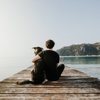 Ein Mann sitzt mit einem Hund auf einem Steg und schaut aufs Wasser. | © AdobeStock-327201693