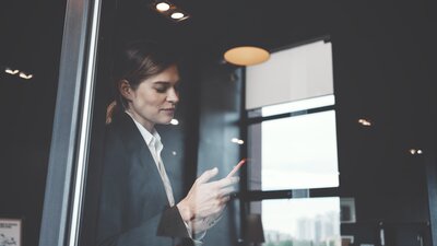 Eien Frau mit Smartphone in der Hand steht an einem Fenster eines Bürogebäudes. | © AdobeStock-158431875