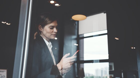 Eien Frau mit Smartphone in der Hand steht an einem Fenster eines Bürogebäudes. | © AdobeStock-158431875