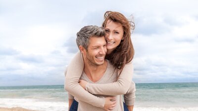 Ein Mann trägt eine Frau Huckepack an einem Strand. | © AdobeStock-61341087
