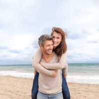 Ein Mann trägt eine Frau Huckepack an einem Strand. | © AdobeStock-61341087