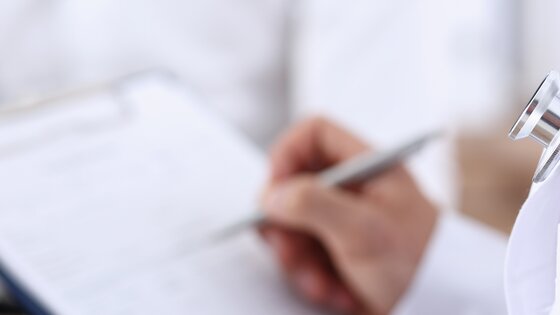 Detailaufnahme eines Arztes mit Stethoskop, der etwas auf einem Klemmbrett notiert. | © AdobeStock-293499926